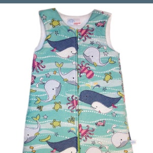 Może przedstawiać: Turkusowy śpiwór z białym zamkiem błyskawicznym. Śpiwór ma wzór białych wielorybów, różowych ośmiornic, zielonych żółwi morskich i żółtych gwiazd morskich na turkusowym tle.