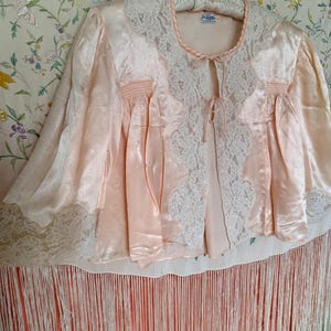 May include: A pale peach satin bed jacket featuring white lace trim along the collar, front opening, and sleeve cuffs. The garment includes two thin satin ties at the neckline and a long, flowing fringe hemline that extends below the main body.