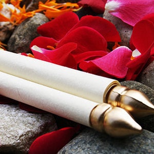 Puede incluir: Un rollo blanco atado con una borla dorada, descansando sobre un lecho de pétalos de flores rojas y naranjas y piedras grises.