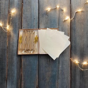 May include: A collection of gold-coloured invitation scrolls with tassels and blank paper inserts, displayed in an open rectangular box. The box sits on a weathered, blue-grey wooden surface. String lights with small bulbs are arranged around the box.