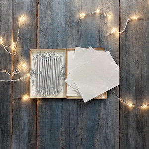 May include: A set of silver pens and white paper in an open box, set on a weathered wooden surface. The pens have decorative tops and are tied with a silver ribbon. Fairy lights are strung around the box.
