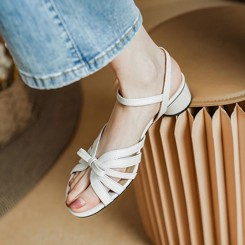 May include: White leather sandals with a bow detail on the toe and a small block heel. The sandals have multiple straps across the foot and an ankle strap. The heel is approximately 1.5 inches.