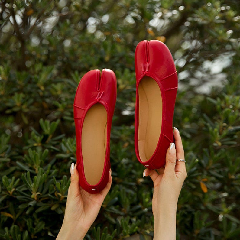 May include: A pair of bright red ballet flats with a split-toe design, held up against a backdrop of green foliage. The shoes have a smooth, glossy finish and a tan interior.