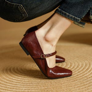 May include: Burgundy leather Mary Jane flats with a square toe and a small strap with a gold buckle. The shoes have a low heel and a glossy finish. The image shows the shoes on a woven surface.