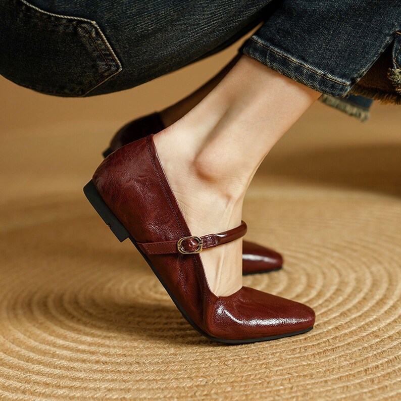 May include: Burgundy leather Mary Jane flats with a square toe and a small strap with a gold buckle. The shoes have a low heel and a glossy finish. The image shows the shoes on a woven surface.