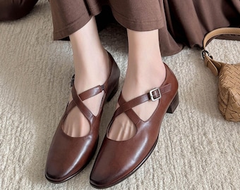 May include: A pair of brown leather Mary Jane style shoes with a criss-cross strap design and a small buckle closure. The shoes have a low heel and a rounded toe. The shoes are a rich, dark brown color.