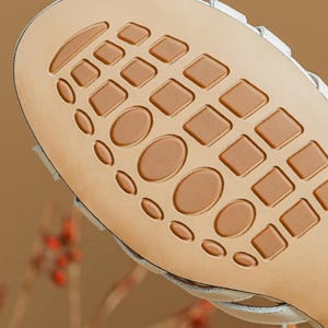 May include: Close-up of the sole of a white sandal. The sole is tan with a pattern of squares and ovals. The sandal's white upper is visible along the edges.
