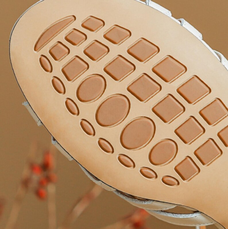 May include: Close-up of the sole of a white sandal. The sole is tan with a pattern of squares and ovals. The sandal's white upper is visible along the edges.