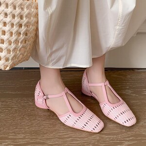 May include: Pink woven flats with a T-strap design. These shoes feature a square toe and a small heel. The flats have a woven texture and a buckle closure on the ankle strap. The shoes are shown on a wooden floor.