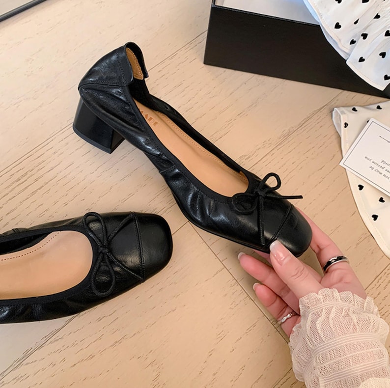 Peut inclure: Ballerines en cuir noir avec un petit talon bloc. Les chaussures ont un bout carr&eacute; et un petit n&oelig;ud sur le devant. L'int&eacute;rieur est de couleur beige clair. Les chaussures sont sur une surface &agrave; grain de bois clair.