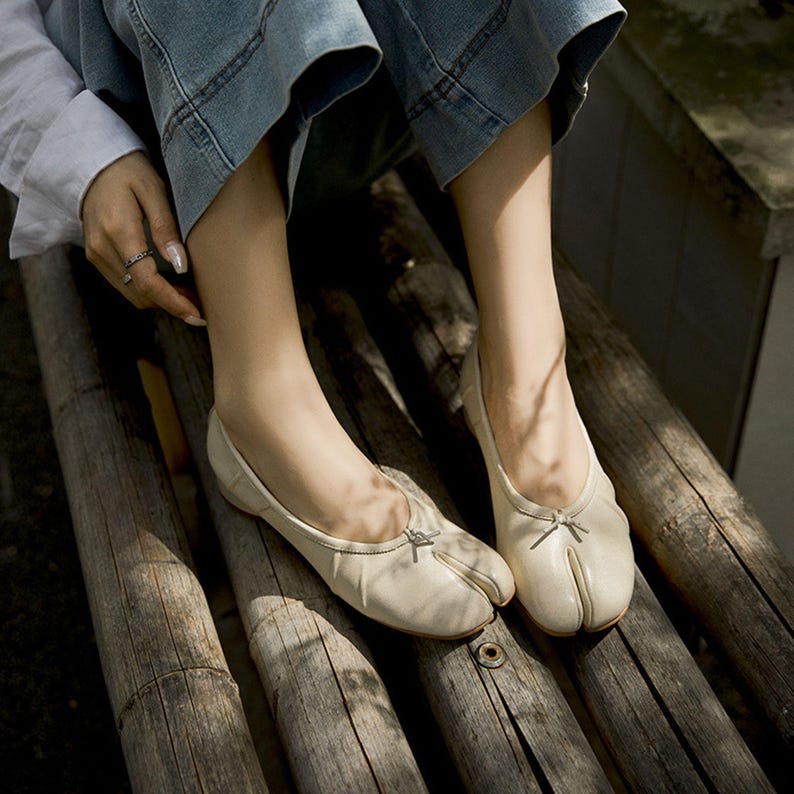 May include: Cream-colored split-toe ballet flats with a small bow detail. The shoes are made of a soft material and have a minimalist design. The flats are shown on a wooden bench, suggesting a casual, everyday style.