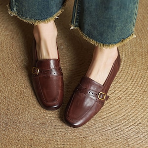 Puede incluir: Un par de mocasines de cuero marr&oacute;n con punta cuadrada y una correa decorativa con hebilla dorada. Los zapatos son usados por una persona que lleva vaqueros azules.