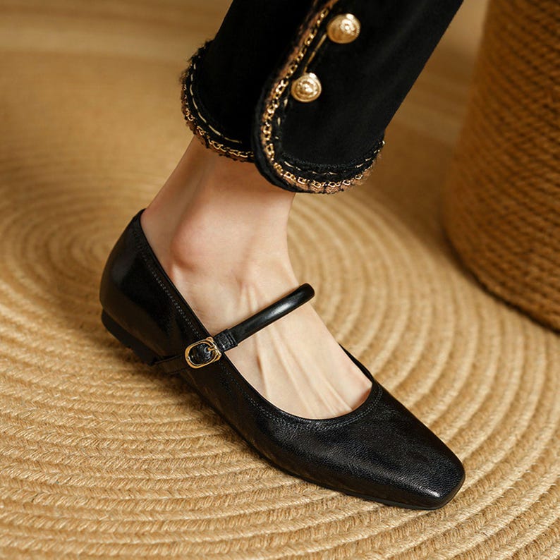 May include: Black leather Mary Jane flats with a square toe and a strap across the instep. The shoes have a small heel and a gold-colored buckle. The flats are on a woven surface.