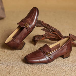 Puede incluir: Un par de mocasines de cuero marr&oacute;n con tac&oacute;n bajo. Los zapatos presentan una puntera cuadrada, una correa en la parte superior con una hebilla dorada y un tac&oacute;n apilado. Los zapatos se exhiben sobre una superficie de tono neutro.
