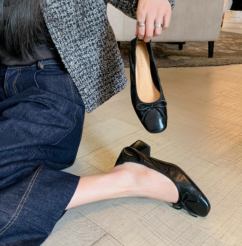 Peut inclure: Ballerines en cuir noir avec un petit n&oelig;ud sur le bout. Les chaussures ont un bout carr&eacute; et un petit talon. L'image montre une chaussure tenue et une autre sur le pied d'une personne.