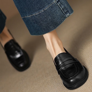 May include: Close-up of black leather loafers with a textured finish. The shoes have a classic design with a rounded toe and a strap detail across the top. The image also shows denim jeans.