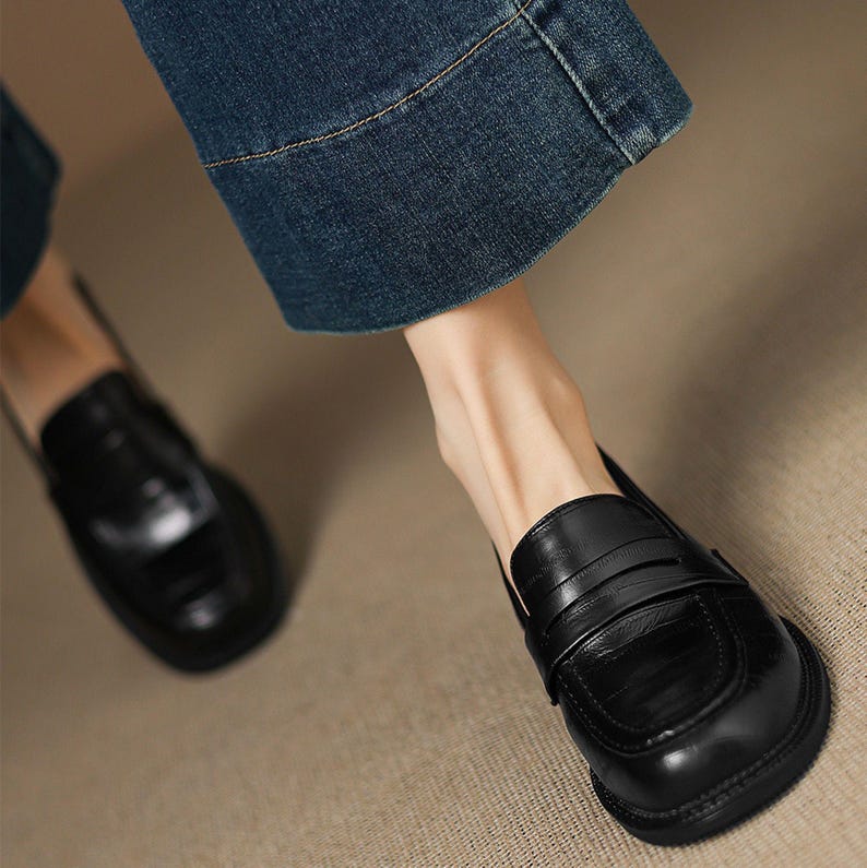May include: Close-up of black leather loafers with a textured finish. The shoes have a classic design with a rounded toe and a strap detail across the top. The image also shows denim jeans.