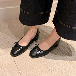 Peut inclure: Une paire de ballerines en cuir noir avec un bout carr&eacute; et un petit n&oelig;ud. Les chaussures ont un petit talon et sont pr&eacute;sent&eacute;es sur un sol en bois clair. Les chaussures sont port&eacute;es avec un pantalon en jean fonc&eacute;.