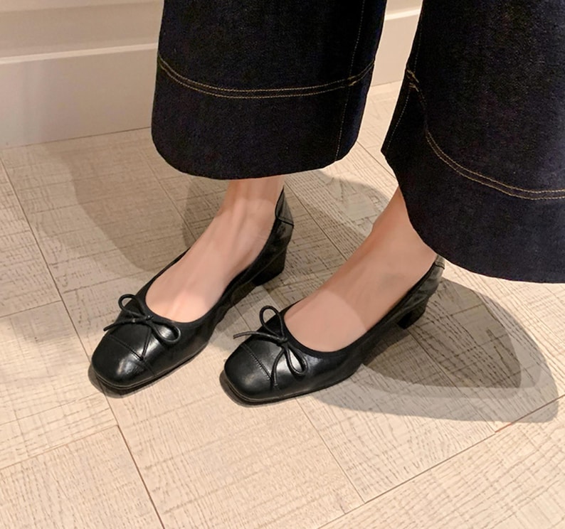 Peut inclure: Une paire de ballerines en cuir noir avec un bout carr&eacute; et un petit n&oelig;ud. Les chaussures ont un petit talon et sont pr&eacute;sent&eacute;es sur un sol en bois clair. Les chaussures sont port&eacute;es avec un pantalon en jean fonc&eacute;.