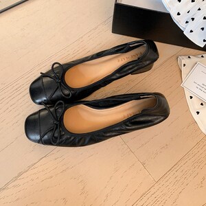 Peut inclure: Une paire de ballerines en cuir noir avec de petits n&oelig;uds et des bouts carr&eacute;s. Les chaussures ont un petit talon et sont pr&eacute;sent&eacute;es sur une surface en bois clair. Le mot "KANEMADE" est visible sur la semelle int&eacute;rieure.