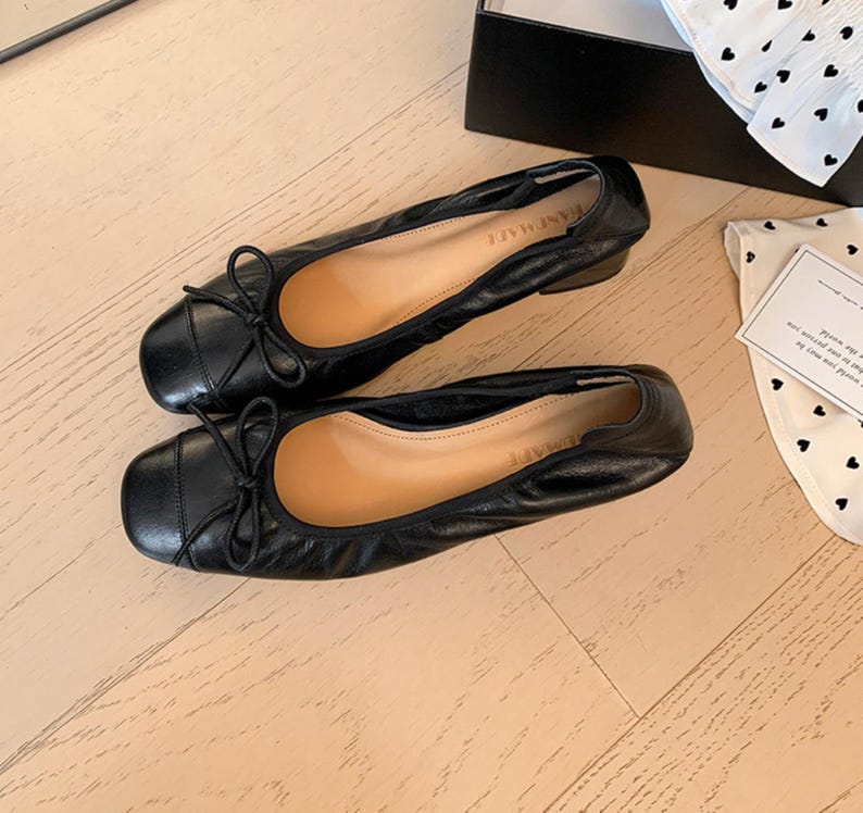 Peut inclure: Une paire de ballerines en cuir noir avec de petits n&oelig;uds et des bouts carr&eacute;s. Les chaussures ont un petit talon et sont pr&eacute;sent&eacute;es sur une surface en bois clair. Le mot "KANEMADE" est visible sur la semelle int&eacute;rieure.