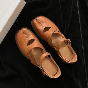 May include: Brown leather sandals with a split-toe design. The sandals have a strap that buckles around the ankle and a textured surface. The shoes are set against a dark background.