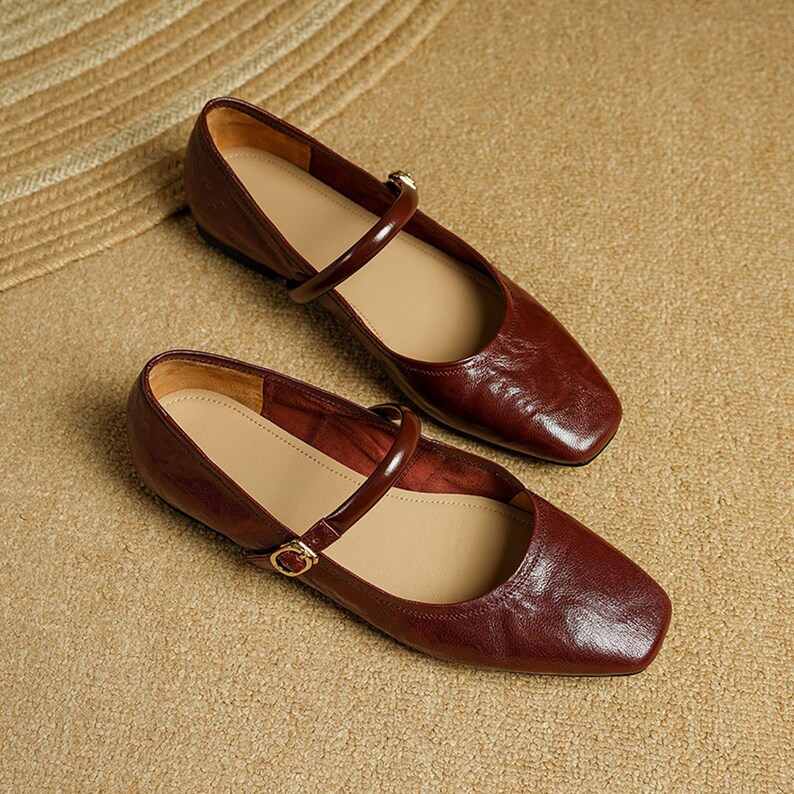 May include: A pair of burgundy leather Mary Jane flats with a square toe and a strap across the top. The shoes have a gold-colored buckle and a light tan insole. The shoes are displayed on a textured, tan surface.