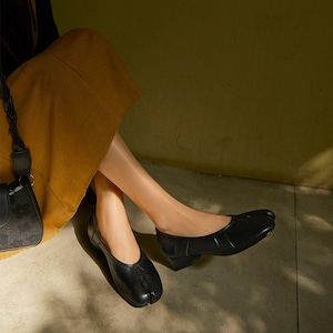 May include: Black leather split-toe flats with a low heel. The shoes have a rounded toe and a strap detail across the top. The design is reminiscent of traditional Japanese footwear. The shoes are worn with a mustard-colored skirt.