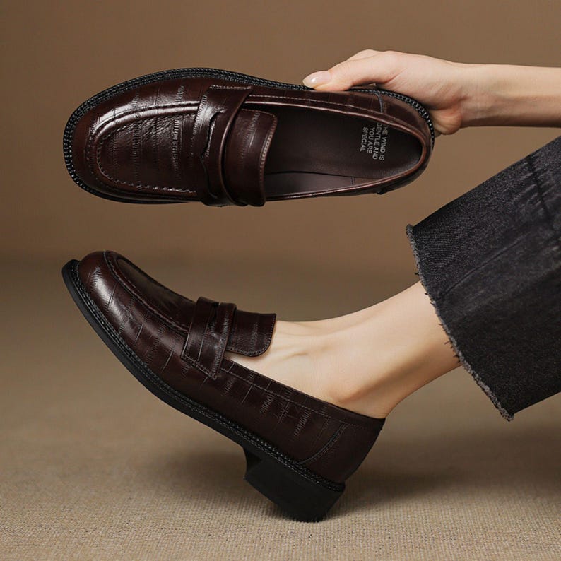 May include: Dark brown leather loafers with a textured finish. The shoes have a classic design with a rounded toe, a strap across the vamp, and a small heel. The sole is black. The image shows one shoe being held and another on a foot.