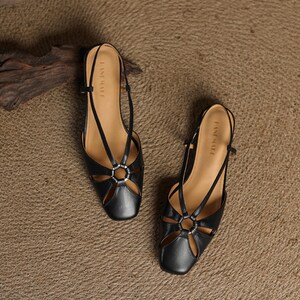 May include: A pair of black leather sandals with a square toe and cut-out design. The sandals feature a silver ring detail and thin straps. The brand name "EASE MATE" is printed on the insole. The sandals are displayed on a textured, neutral-toned surface.