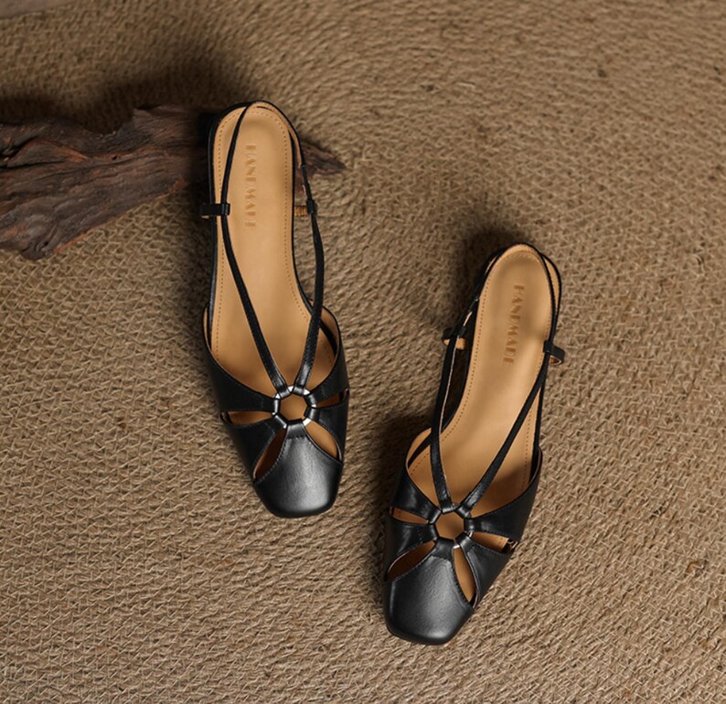May include: A pair of black leather sandals with a square toe and cut-out design. The sandals feature a silver ring detail and thin straps. The brand name "EASE MATE" is printed on the insole. The sandals are displayed on a textured, neutral-toned surface.
