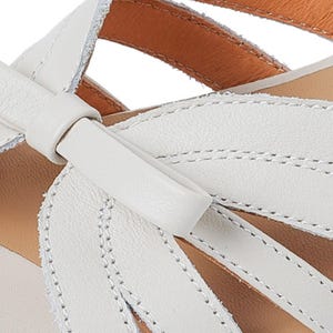 May include: Close-up of a white leather sandal with a bow detail. The sandal features a stitched design and a tan insole. The leather appears soft and the stitching is precise, highlighting the craftsmanship of the footwear.