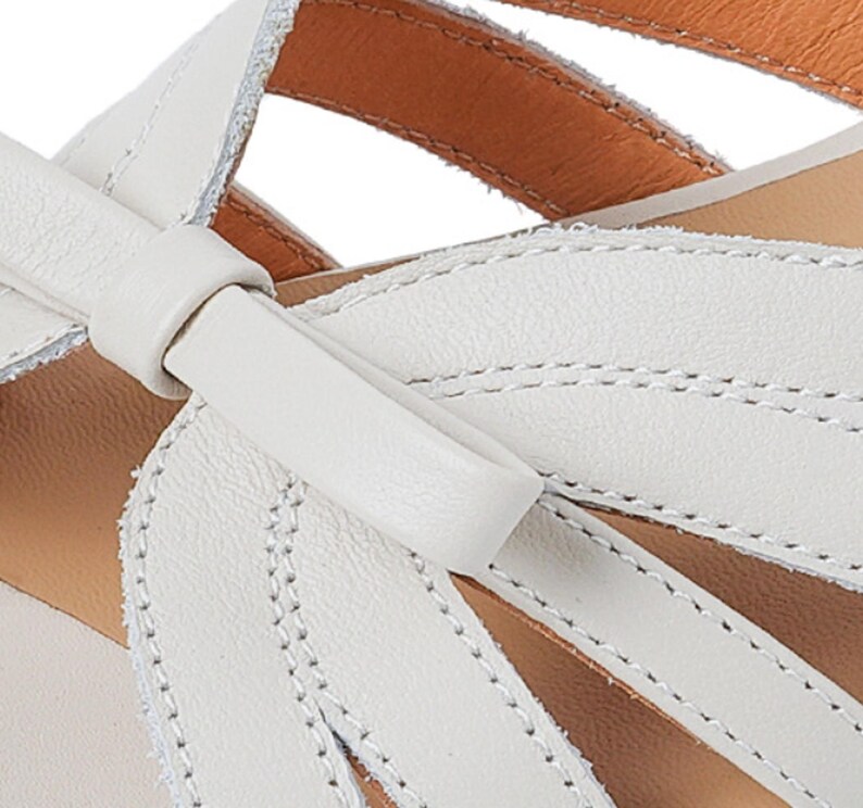 May include: Close-up of a white leather sandal with a bow detail. The sandal features a stitched design and a tan insole. The leather appears soft and the stitching is precise, highlighting the craftsmanship of the footwear.