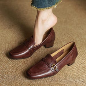 Puede incluir: Mocasines de cuero marr&oacute;n con punta cuadrada y tac&oacute;n peque&ntilde;o. Los zapatos tienen una correa en la parte superior con una hebilla dorada. El cuero tiene un ligero brillo. Los zapatos est&aacute;n sobre una superficie tejida.