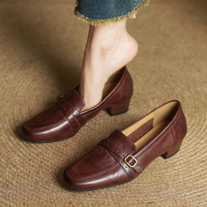 Puede incluir: Mocasines de cuero marr&oacute;n con punta cuadrada y tac&oacute;n peque&ntilde;o. Los zapatos tienen una correa en la parte superior con una hebilla dorada. El cuero tiene un ligero brillo. Los zapatos est&aacute;n sobre una superficie tejida.