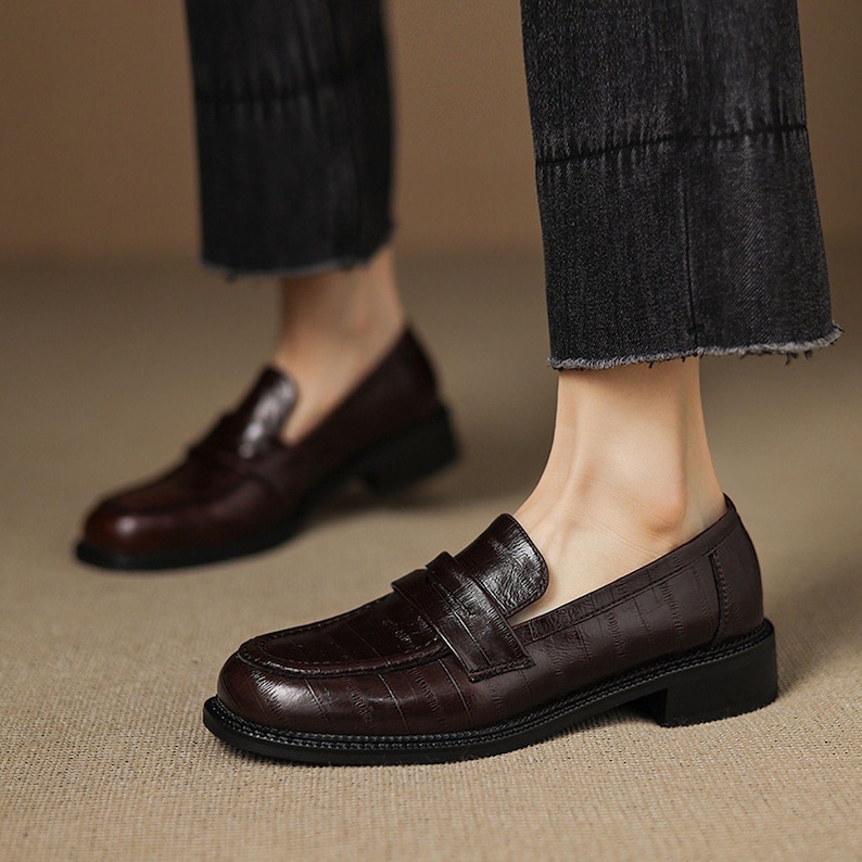 May include: Dark brown leather loafers with a textured, embossed pattern. The shoes have a classic penny loafer design with a strap across the top and a low, block heel. The soles are black.