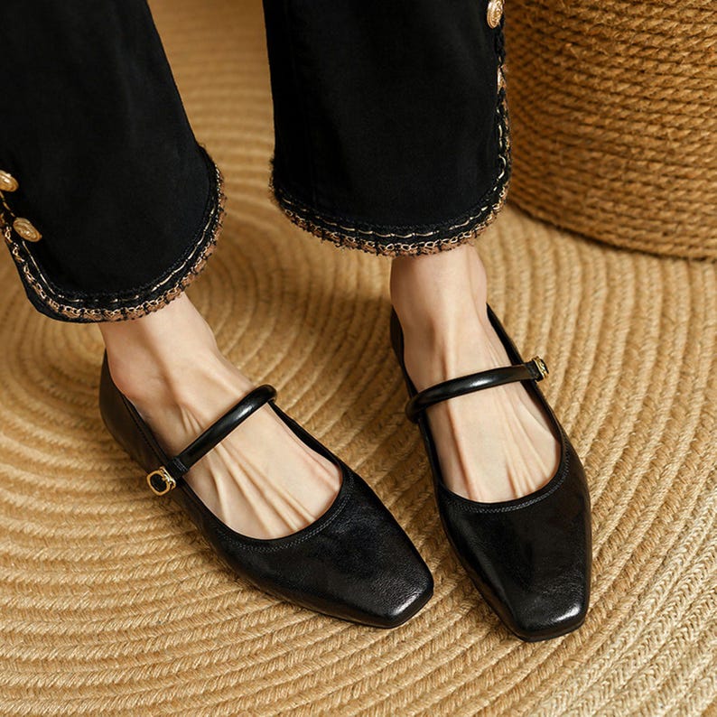 May include: Black leather Mary Jane flats with a square toe and a strap across the top of the foot. The shoes have a small gold buckle and are worn with black pants.