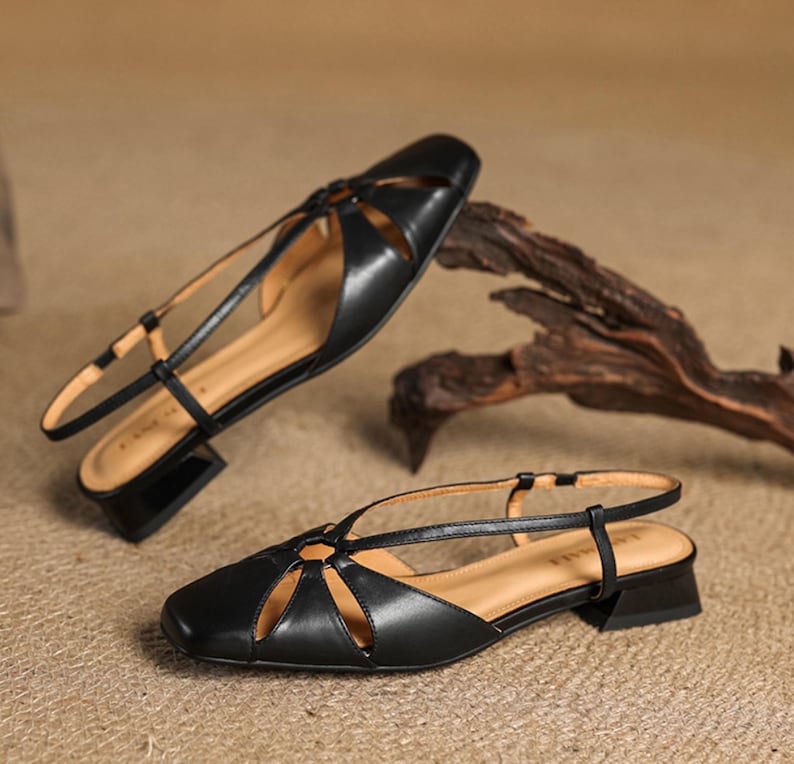 May include: A pair of black leather sandals with a square toe and a low, block heel. The sandals feature cut-out details on the vamp and a thin ankle strap. The interior is a light tan color.
