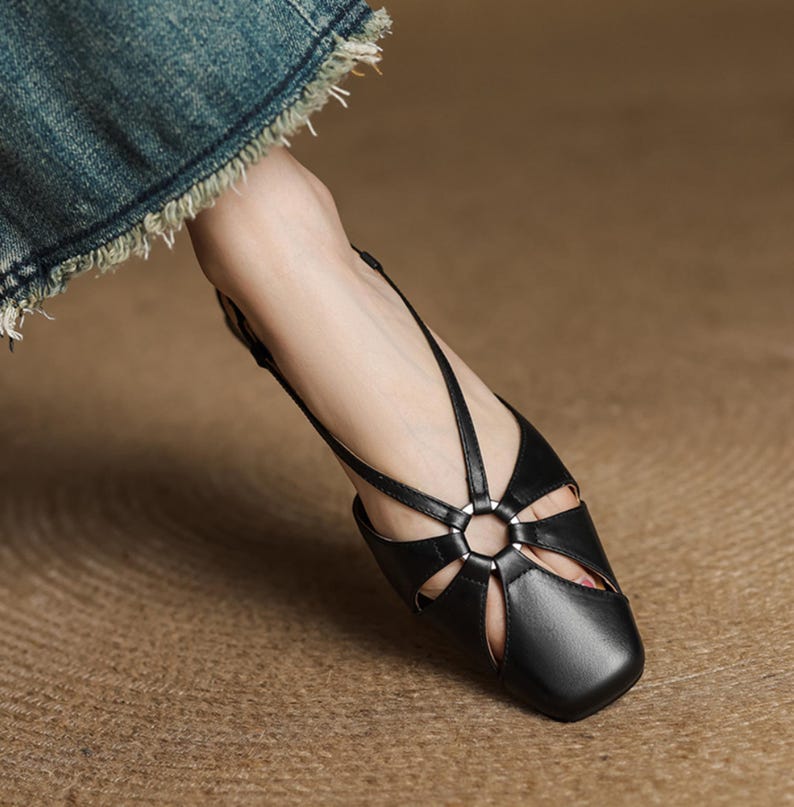 May include: Black leather sandal with a square toe and cut-out design. The sandal features a slingback strap and a decorative metal ring at the center. The shoe is shown on a textured surface.