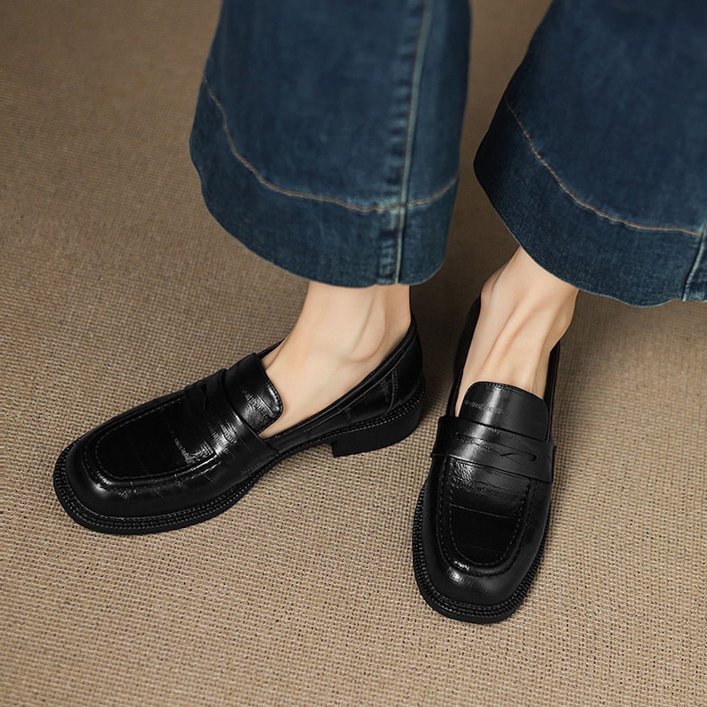 May include: A pair of black leather loafers with a square toe and a low heel. The shoes have a textured finish and a strap detail across the top. The loafers are worn with blue denim jeans.