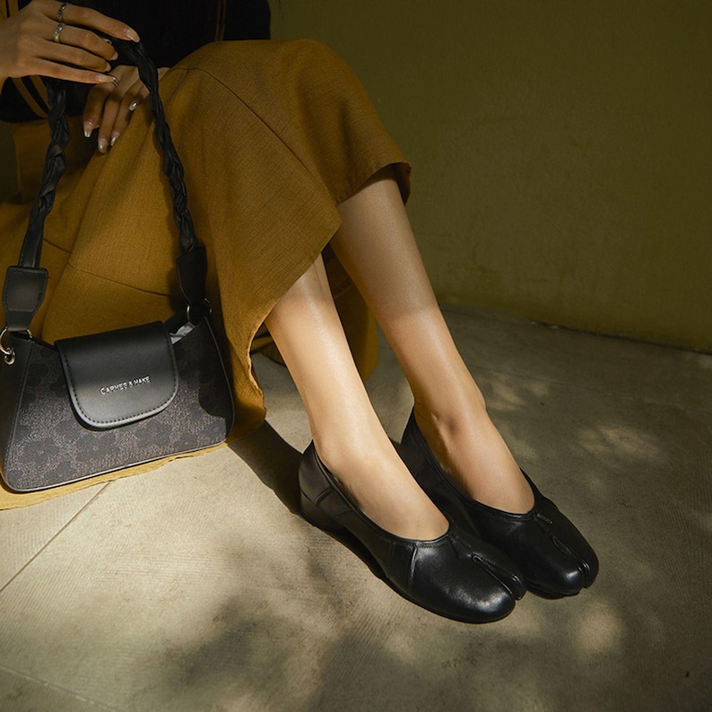 May include: Black leather ballet flats with a split-toe design. The shoes are paired with a black handbag with a patterned design and the text "CHARLES & KEITH". The bag has a braided strap. The person is wearing a mustard-colored skirt.