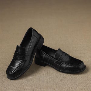 May include: A pair of black leather loafers with a textured finish. The shoes have a classic slip-on design with a strap across the top and a low heel. The loafers are displayed on a neutral background.