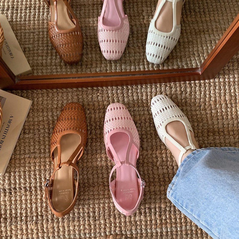 May include: An assortment of woven sandals in various colors. The selection includes brown, pink, and white open-toe flats and sandals. The shoes are displayed on a woven surface, with a mirror reflecting some of the footwear.