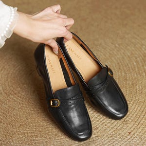 Puede incluir: Mocasines de cuero negro con punta cuadrada y hebilla dorada. Los zapatos tienen una correa en la parte superior y se exhiben sobre una superficie tejida. El interior del zapato es de color beige.