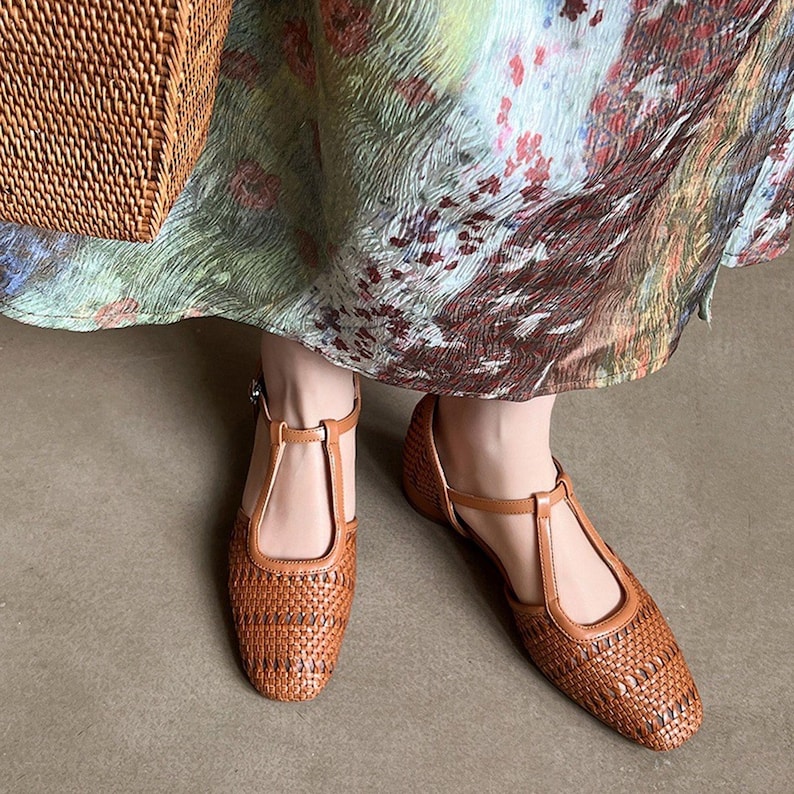 May include: A pair of brown woven leather flats with a T-strap design. The shoes have a square toe and a buckle closure. The flats are worn with a floral print dress.