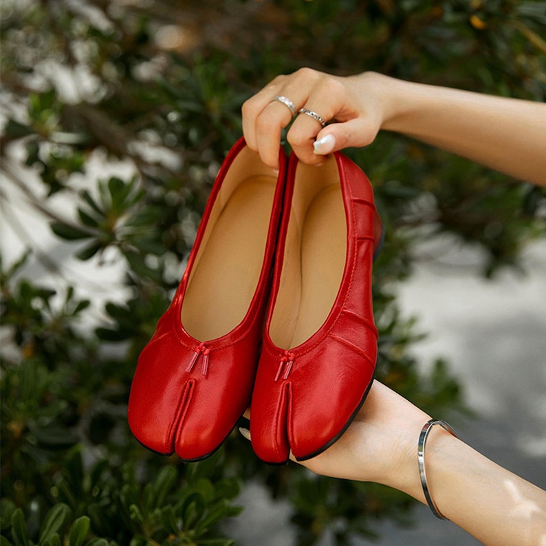 May include: A pair of bright red ballet flats with a split-toe design. The shoes have a smooth, leather-like texture and a small bow detail at the top. The interior is a light tan color.