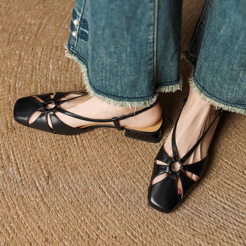 May include: Black leather sandals with a square toe and cut-out design. The sandals have a thin ankle strap and a small, low heel. The shoes are worn with blue denim jeans with frayed edges.