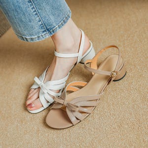 May include: Two pairs of sandals. One pair is white with a bow detail, the other is tan with a bow detail and a small heel. Both sandals have straps and are designed for warm weather.