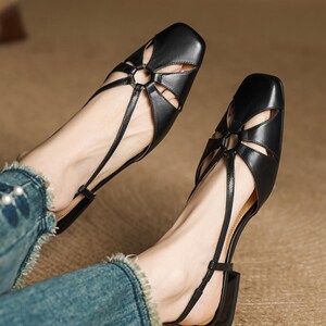 May include: Black leather sandals with a square toe and a floral-shaped cutout design on the vamp. The sandals have thin straps and a small block heel. The shoes are worn with blue jeans.
