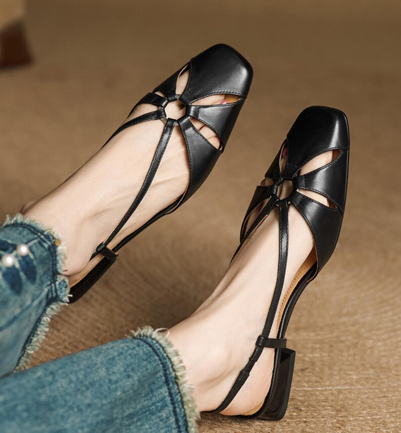 May include: Black leather sandals with a square toe and a floral-shaped cutout design on the vamp. The sandals have thin straps and a small block heel. The shoes are worn with blue jeans.